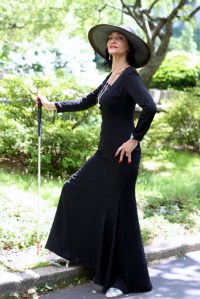 woman in amazing black dress, giant hat and silver jewelry stands jauntily with a white cane